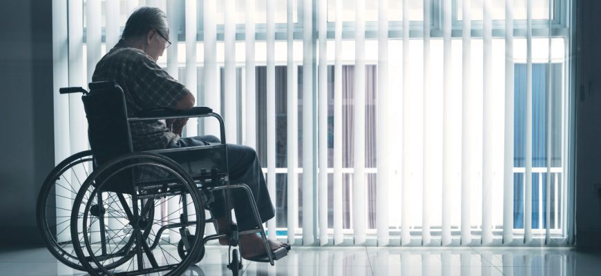 Lonely senior resident in assisted living facility looks out the window solemnly as he sits in his wheelchair.