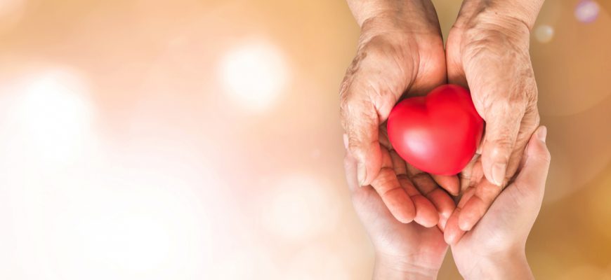 Two pairs of hands, one older and one younger, cup a small red heart which symbolized heart and cholesterol health in senior citizens.